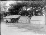 Five unidentified men in cloth caps and two boys in front of a large tent with an awning with 'Raukura' camp sign, next to two smaller tents, tall trees beyond, [Sumner?], Christchurch