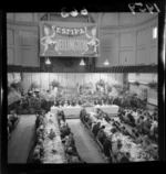 Festival of Wellington dinner in the Wellington Town Hall, showing unidentified guests sitting at long tables, and musicians performing on a stage covered with a floral display