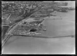 Mechanics Bay, Auckland City, showing Auckland Harbour Wharf area with TEAL (Tasman Empire Airways Limited) Air Base with hanger and flying boats on land and harbour, Tamaki Drive and Quay Street, view west to ships docking, rail yards and City Centre beyond