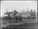 Christchurch Railway Station, draped in flags and garlands of ferns and leaves, people milling around with horse drawn carriages, with 'A Merry Christmas To You All'