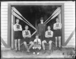 Unidentified men's coxed four rowing team with a young boy coxswain and coach, with crossed oars behind, outside the Avon Rowing Club boatshed, Christchurch