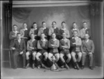 Studio portrait, unidentified men's hockey team and coaches, with hockey sticks and large trophy cup with lid and small shield plaques around its base, Christchurch