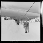 An unidentified woman on a chairlift at the New Zealand Ski Championship at Mt Ruapehu, showing skiers and spectators in the background