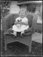 A very young girl sitting in a cane chair in the backyard, holding a porcelain dog, Hawke's Bay District