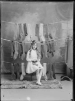 Studio portrait of an unidentified girl sitting on a wooden stool knitting a sock, with three rows of socks hanging behind her, possibly Christchurch district