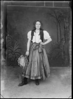 In front of a false backdrop, portrait of an unidentified young woman in a gypsy costume, in a long pleated skirt with metal spheres, a white blouse with a dark vest, necklace, bracelets and a headband with metal spheres, standing holding a scarf and tambourine with streamers, probably Christchurch region