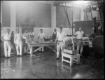 An interior view of Heretaunga Co-op Dairy Co Ltd, showing a group of unidentified men working in the factory, a man standing next to a butter processing machine, a man standing next to scales with some butter on it, and a group of men standing next to a packing table with some packing boxes on it, Heretaunga Plains, Hawkes Bay