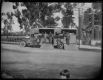 A side view of a two horse accommodation horse float truck in front of the Maori gateway entrance to Ebbett Park, Hastings, showing a group of unidentified men standing in front of the truck and two horses in the stalls and an unidentified girl in the foreground