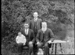 Outdoors portrait of three unidentified men with baby in christening gown and a small pug dog in front, probably Christchurch region