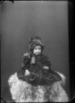 Studio Slater family portrait of a toddler girl in a dark patterned dress with large collar and bow with matching pleated hat with ribbon sitting on fur rug, Christchurch
