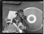 Portrait of Squadron Leader D Greig in flight gear standing beside his plane, Ardmore Aerodrome, Auckland Region
