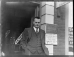 Mr SB Taylor, public accountant and auditor, and secretary of Gore Aero Club, standing at the entrance to his office, Gore, Southland Region