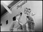 Bristol Freighter tour, Mr W R Burns, receives poi brooch from Guide Emily, who is wearing a Maori cloak and a head scarf, standing by a Bristol Freighter aircraft, [Rotorua?]