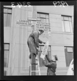 Two men from the Automobile association, erecting roadsigns on a power pole on Oriental Parade, Wellington, indicating direction and distances of various suburbs