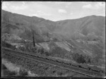 Distant view of a train ascending the Rimutaka Incline, looking across a narrow gully, with track showing the centre rail for the Fell system, in foreground.