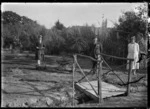 Albert Percy Godber standing by a footbridge over a narrow creek. His daughter Phyllis stands on the left, with a lawn-mower, and his wife Laura is on the right, in their garden at Silverstream