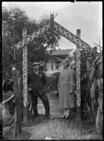 Albert Percy Godber and his wife Laura Godber standing under an archway decorated in Maori style in their garden at Silverstream