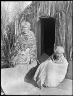 Two elderly Maori with blankets and piupiu on a woven mat outside a raupo whare