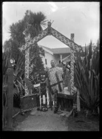 Albert Percy Godber wearing a Maori cloak, holding a taiaha, standing beside a gateway with Maori carvings and designs, working on a Maori carving, 1932