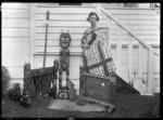Phyllis Mary Godber wearing a Maori cloak, holding a taiaha, standing beside a collection of Maori carvings, including two fire-screens, carved by her father Albert Percy Godber