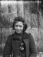 Unidentified Maori girl standing in front of a raupo whare at Karaka Bay, Wellington