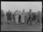 Golfers Bobby Locke (South Africa) and Bob Charles (New Zealand) holding golf clubs and walking on the Miramar Golf Course surrounded by a crowd dressed for windy weather