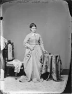Miss Wilkie, wearing a bvery long skirt and bodice, and holding a whip and resting a hand on the book on a table