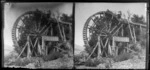 Group of unidentified women and children sitting at the base of a large wooden wheel at a mill, Kinloch, Taupo District