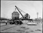 Men using a crane to load a tram for Whanganui, onto a train carriage, Christchurch railyards