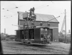 Men in a Christchurch Tramways vehicle, repairing overhead tram wires, including tram shed in background