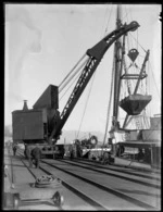 Wharf area, Westport, with crane on rails lifting coal hopper, ship and Westport Harbour beyond