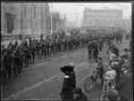 View of a procession of World War I soldiers marching through Cathedral Square, Christchurch, with crowds of people looking on, commercial building beyond 'Barnett & Co, Chemists'