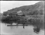 The Landing, Taumarunui, with an unidentified Maori man in a waka on the [Wanganui?] River, and a dwelling, with wooden walls and roof on the bank behind