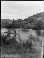The Landing, Taumarunui, an unidentified Maori man in a waka on the [Wanganui?] River, and a dwelling, with wooden walls and roof on the bank behind