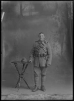 Studio portrait of unidentified World War One soldier with bandolier over left shoulder and ammunition waist belt with revolver holster wearing stirrups standing with hat with silver fern badge, Christchurch