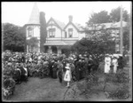 Staff and students from Christ's College, Christchurch, with family members in front of a two storey wooden house