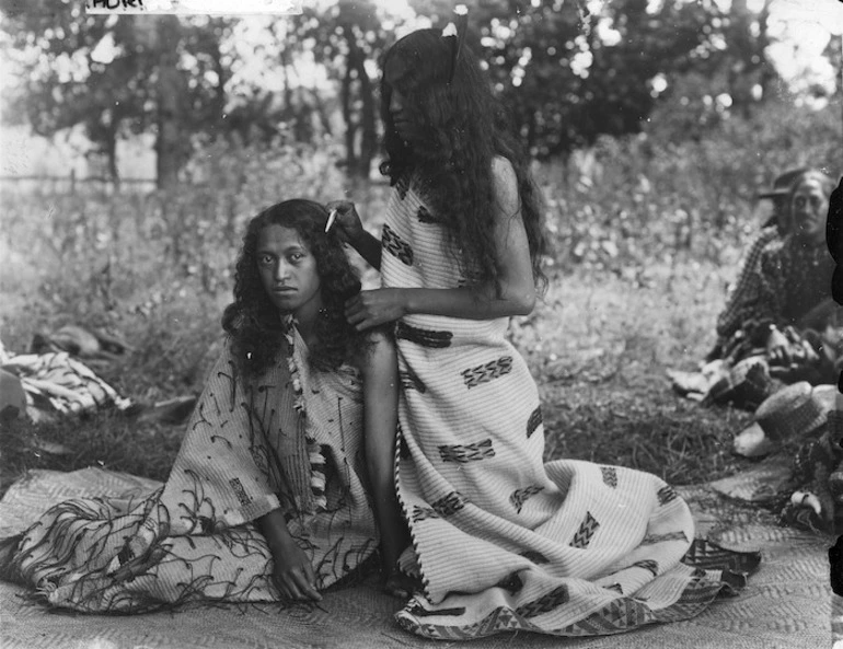Image: A young woman, wearing a Maori cloak, combing the hair of another