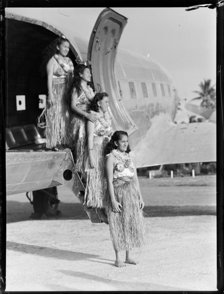 Image: Four unidentified local girls wearing hula skirts standing on a C47 transport aircraft ladder into cargo door, Rarotonga airfield, Cook Islands