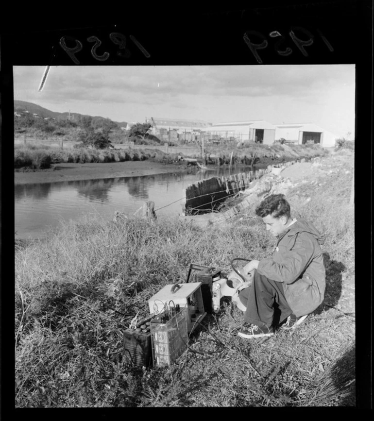 Image: A technician of the Institute of Nuclear Sciences studying recording equipment by the Waiwhetu stream, Lower Hutt