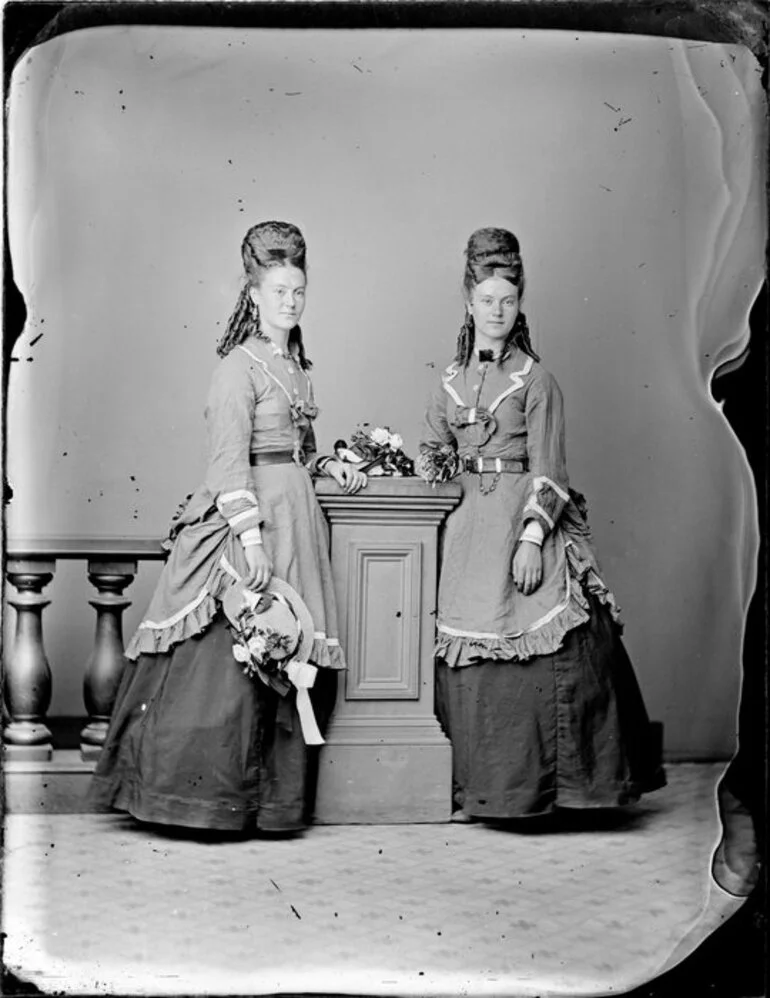 Image: Two unidentified young women, wearing matching full length skirts and elaborate overdresses, with identical hairstyles, featuring ringlets and padded upswept hair