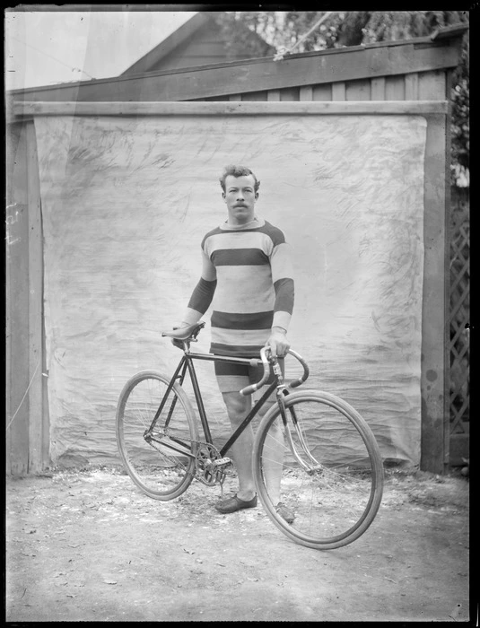 Unidentified man in cycling attire standing with his road racing bicycle within a backyard area with wooden fence behind, probably Christchurch region