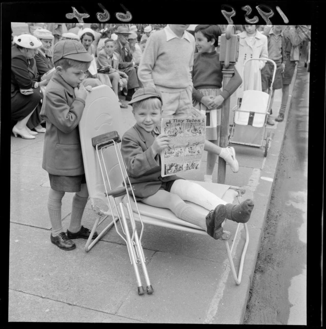 Spectators at a Wellington parade, including two unidentified small boys in school uniform, one with a broken leg in a plaster cast, who is sitting in a folding chair and reading Jack and Jill childrens' magazine