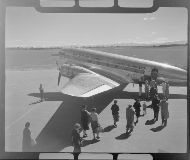 Harewood, Christchurch, New Zealand National Airways Corporation DC3 on runway, with passengers embarking through rear doorway of plane via stairs, snow covered mountains beyond
