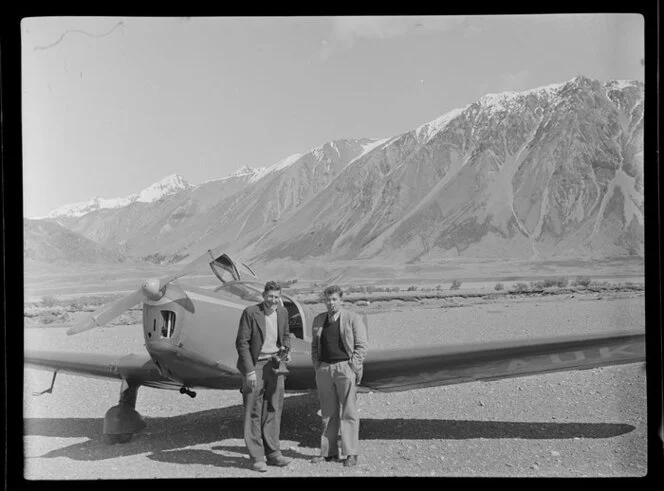 Lake Ohau, Central Otago, view of shingle river bed with two men (one with camera) standing beside a Mount Cook Whitley Straight (ZK AUK) tourist aircraft, snow capped mountains beyond