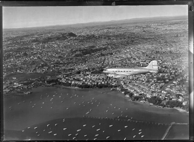 New Zealand National Airways Corporation (NAC) Flagship Dakota aircraft in flight over Auckland, showing boats in the harbour