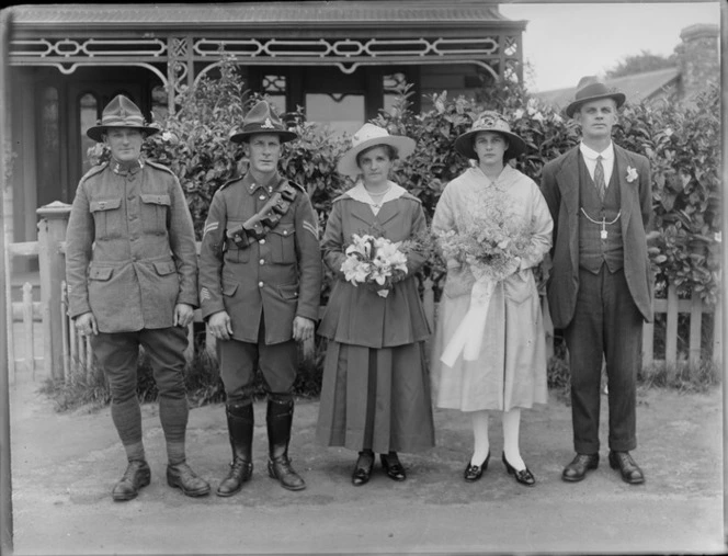 Unidentified wedding party portrait in front of a house with veranda, two men in army uniform, women holding flowers, probably Christchurch region