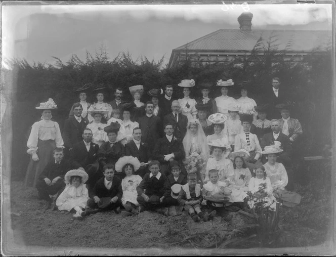 Wedding group portrait in front of a tall hedge with house beyond, unidentified bride with long veil and groom with extended family and children, women with hats, probably Christchurch region