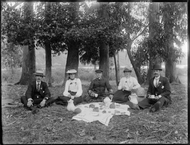 Group of unidentified young men and women with hats, one man in uniform, having a picnic in front of tall trees, with a bay and steep hills beyond, probably Christchurch region