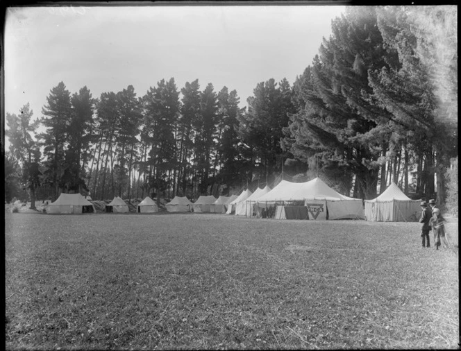 Eleven large double pole top tents at the end of a large field under tall trees, one tent with YMCA name and symbol on it, two men and a child foreground, Christchurch