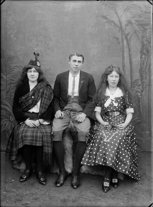 Portrait of an unidentified [performance?] group, showig two women and a man, with a painted backdrop, one woman wearing Scottish national costume including tartan skirt and sash and the other woman wearing a poker dot dress, embroidered with a harp, the man in knickerbockers, probably Christchurch district